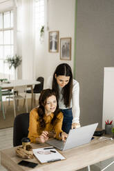 Geschäftsfrau mit Tochter, die auf einen Laptop schaut, während sie einen Keks am Schreibtisch im Heimbüro isst - GIOF11473