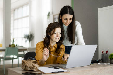 Lächelnde Tochter mit Mutter, die auf einen Laptop schaut, während sie an einem Schreibtisch im Büro zu Hause sitzen - GIOF11474