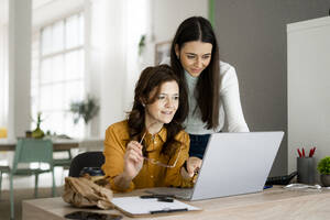 Lächelnde Tochter mit Mutter, die auf einen Laptop schaut, während sie an einem Schreibtisch im Büro zu Hause sitzen - GIOF11474