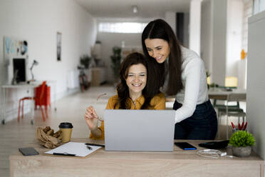 Smiling mother and daughter working on laptop at desk in home office - GIOF11475