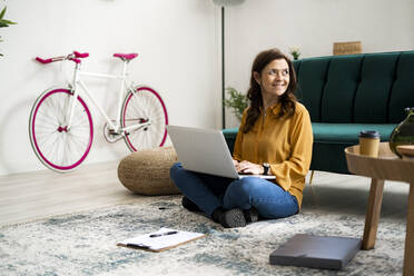 Smiling woman with laptop looking away while sitting on floor at home - GIOF11486