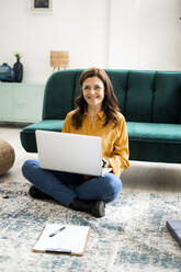 Smiling woman with laptop sitting on floor at sofa in living room - GIOF11487