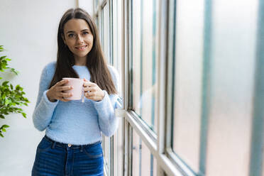 Lächelnde junge Frau mit Kaffeetasse, die am Fenster im Wohnzimmer lehnt - GIOF11503