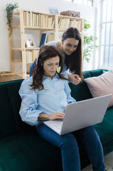 Geschäftsfrau, die einen Laptop benutzt, während eine junge Frau zu Hause auf dem Sofa hilft - GIOF11511