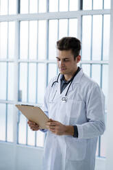 Männlicher Arzt betrachtet Dokumente und hält ein Klemmbrett im Krankenhaus - GIOF11524