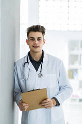 Male medical professional with clipboard leaning on wall in hospital - GIOF11526