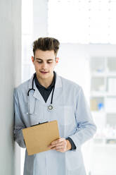 Männlicher Arzt schreibt Notizen, während er sich im Krankenhaus an die Wand lehnt - GIOF11527