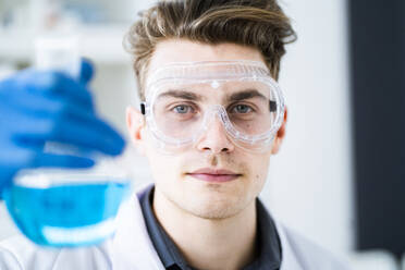 Young scientist wearing protective eyewear while holding flask with chemical in laboratory - GIOF11546