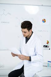 Junger männlicher Wissenschaftler schaut auf eine Tabelle, während er auf einem Schreibtisch im Labor sitzt - GIOF11584