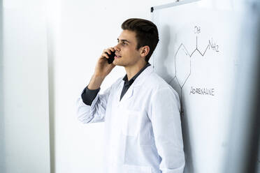 Junger männlicher Wissenschaftler, der mit seinem Smartphone spricht, während er vor einer Tafel im Labor steht - GIOF11587