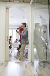 Man having coffee while using smart phone leaning at doorway in office - AFVF08338