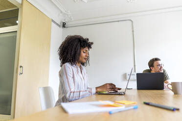 Junge Unternehmerin mit einem männlichen Kollegen, der an einem Laptop arbeitet, während er im Sitzungssaal im Büro sitzt - AFVF08368