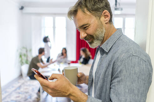 Lächelnder männlicher Unternehmer, der während einer Kaffeepause im Büro ein Smartphone benutzt - AFVF08372