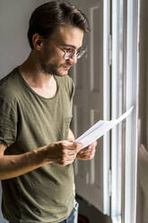 Young male entrepreneur looking at documents while standing near window in office - AFVF08383