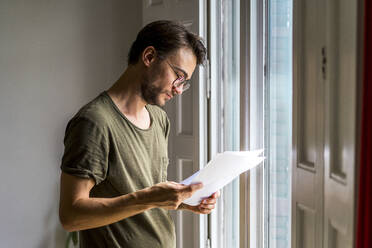 Male entrepreneur going through strategy while standing near window at office - AFVF08384