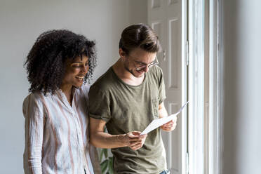 Happy male and female entrepreneurs looking at document while standing near window at office - AFVF08385