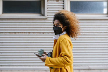 Young woman with coffee cup holding mobile phone while standing by wall during pandemic - EGAF01959
