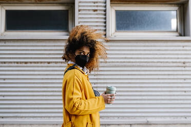 Afro woman with coffee cup standing by wall during COVID-19 - EGAF01960