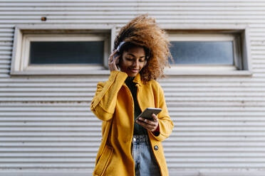 Smiling Afro woman using mobile phone while listening music through headphones against wall - EGAF01965