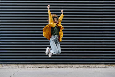 Carefree woman with arms raised jumping against wall - EGAF01970