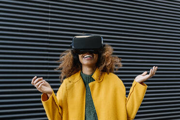 Cheerful Afro woman watching video through virtual reality simulator against wall - EGAF01973