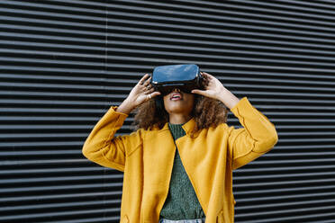 Afro woman in warm clothing watching video through virtual reality simulator against wall - EGAF01974