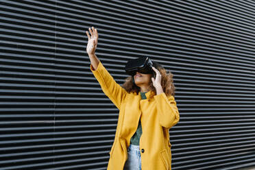 Afro woman with arm raised watching video through virtual reality simulator by wall - EGAF01975