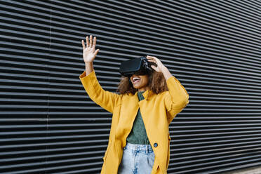 Cheerful Afro woman with arms raised watching video through virtual reality simulator by wall - EGAF01976