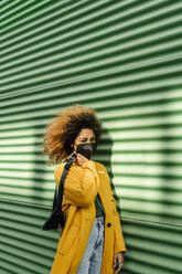 Afro woman with protective face mask holding waist pack by green wall during COVID-19 - EGAF01980