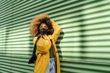 Afro-Frau mit Hand im Haar, die einen Rucksack an der grünen Wand hält, während einer Pandemie - EGAF01981