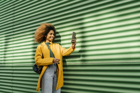 Smiling Afro woman taking selfie through mobile phone while standing by green wall - EGAF01982