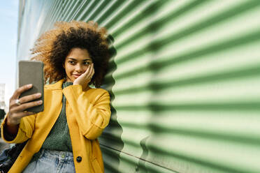 Lächelnde Afro-Frau mit Hand auf der Wange, die ein Selfie mit ihrem Mobiltelefon macht, während sie sich an eine grüne Wand lehnt - EGAF01984