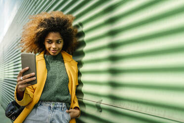Smiling Afro woman with hands in pockets taking selfie through smart phone while leaning on green wall - EGAF01985