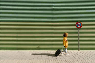 Afro-Frau, die einen Koffer zieht, während sie auf dem Fußweg an der grünen Wand spazieren geht - EGAF01989
