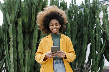 Smiling frizzy haired woman using mobile phone while cactus in background - EGAF01991