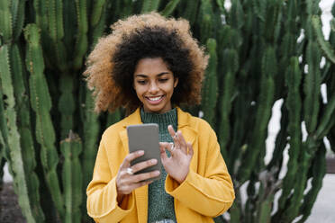 Smiling Afro woman using smart phone while standing against cactus plants - EGAF01993