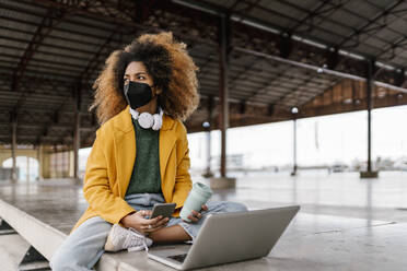 Afro-Frau mit Mobiltelefon und Kaffeetasse, die wegschaut, während sie mit einem Laptop an einer Sitzecke sitzt - EGAF01994