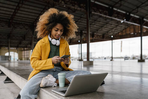 Afro-Frau benutzt ein Smartphone, während sie vor einem Laptop sitzt - EGAF01999