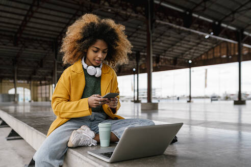 Afro woman using smart phone while sitting in front of laptop - EGAF01999