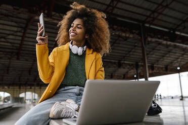 smiling Afro woman using mobile phone while sitting in front of laptop - EGAF02000