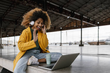 Fröhliche Afro-Frau mit In-Ear-Kopfhörern, die während eines Videogesprächs über einen Laptop im Sitzbereich mit der Hand winkt - EGAF02001