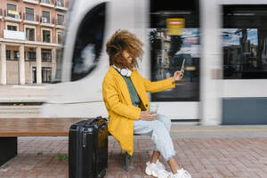 Lächelnde Afro-Frau mit zerzaustem Haar, die ein Selfie mit ihrem Mobiltelefon macht, während sie auf einer Bank an einer Straßenbahnhaltestelle sitzt - EGAF02003