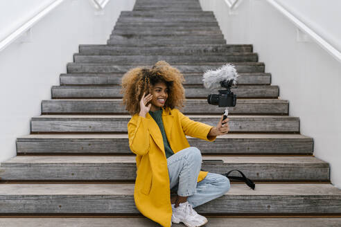 Lächelnde Afro-Frau, die mit der Hand winkt, während sie auf einer Treppe sitzend durch die Kamera vloggt - EGAF02005