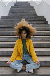 Afro woman with tousled hair meditating while sitting on staircase - EGAF02010