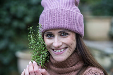 Porträt einer schönen Frau mit rosa Strickmütze, die frischen Rosmarin hält und in die Kamera lächelt, lizenzfreies Stockfoto