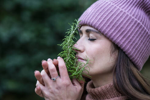 Portrait of beautiful woman wearing pink knit hat smelling fresh rosemary with closed eyes - AKLF00112