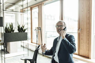 Businessman gesturing while talking on telephone in office corridor - GUSF05292