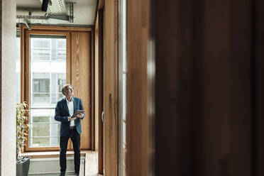 Senior businessman with digital tablet looking up while standing in office corridor - GUSF05295