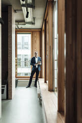 Businessman standing against window in office corridor - GUSF05296