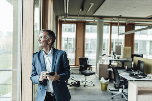 Lächelnder männlicher Unternehmer, der ein Mobiltelefon hält und durch ein Fenster im Büro schaut - GUSF05299